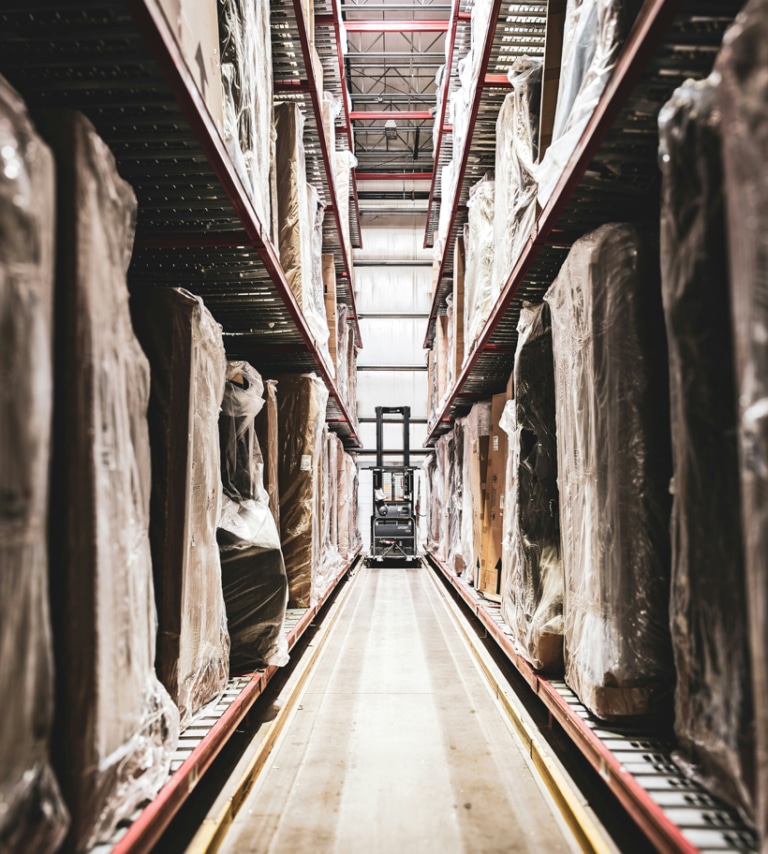 A forklift operates between tall shelves stacked with large, wrapped items in a warehouse aisle.