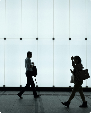 Two people are silhouetted against a large, white, backlit wall; one is walking while holding a bag, and the other is looking at a phone while carrying a purse.