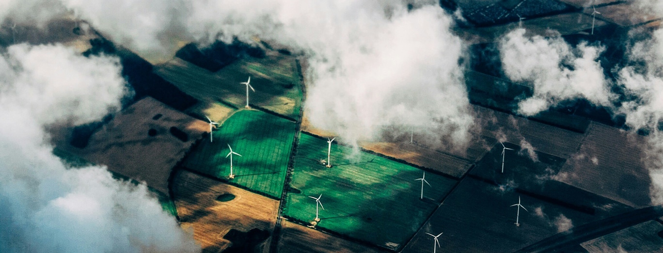 Aerial view of farmland with multiple wind turbines scattered across fields, partially covered by clouds.