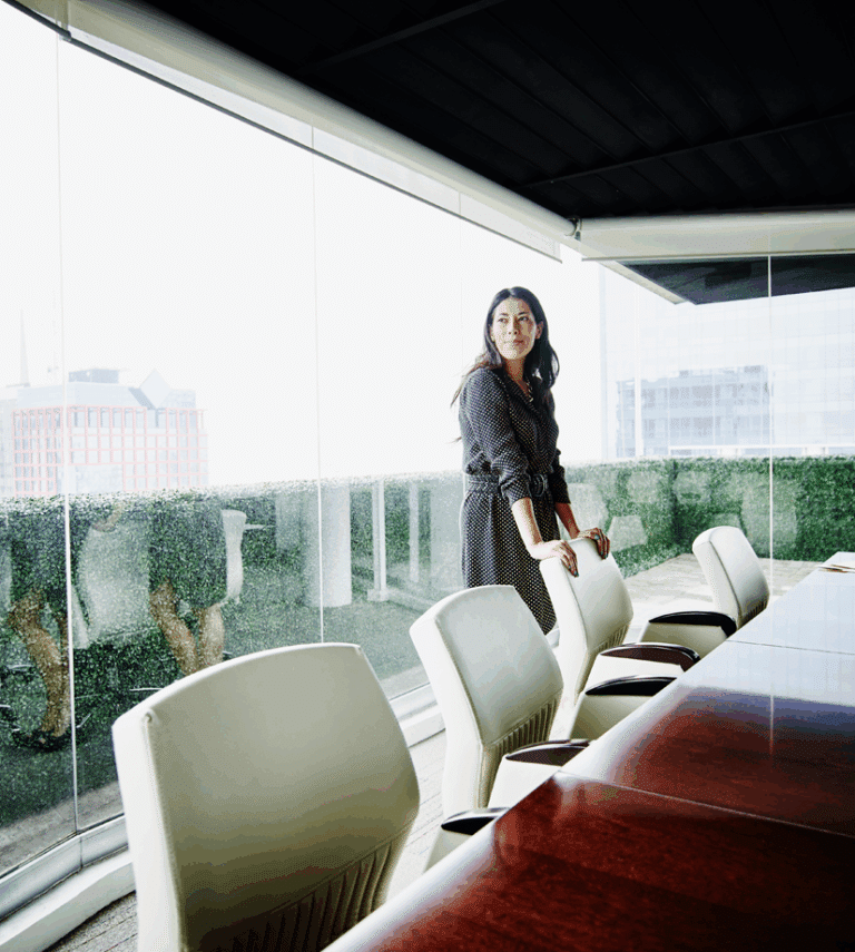 A woman stands at the end of a long conference table in a modern office with large glass windows and city buildings visible outside.