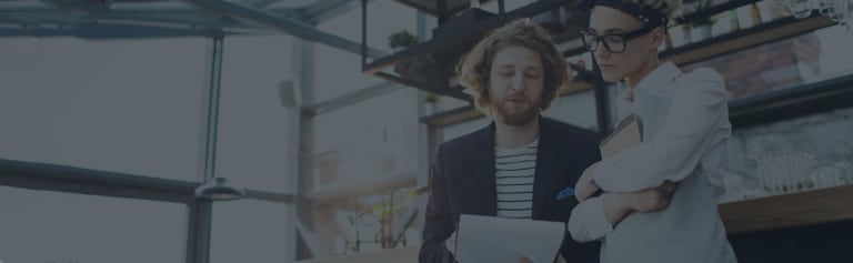 Two people stand indoors reviewing documents; one person holds papers while the other holds a notebook and listens attentively.