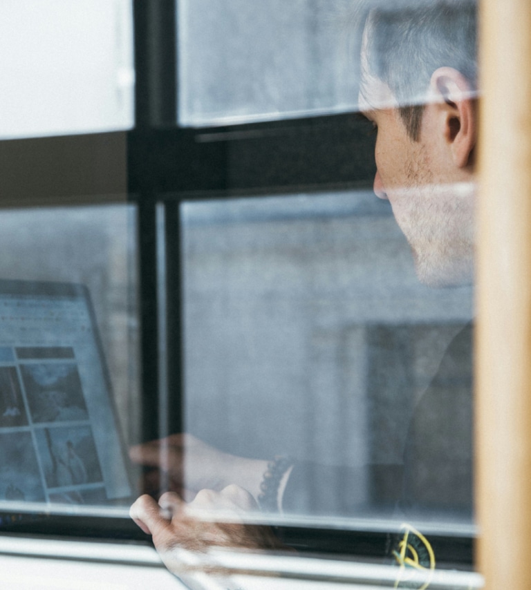 A person is using a laptop indoors, viewed through a window. The screen displays several images and the person is interacting with the device.