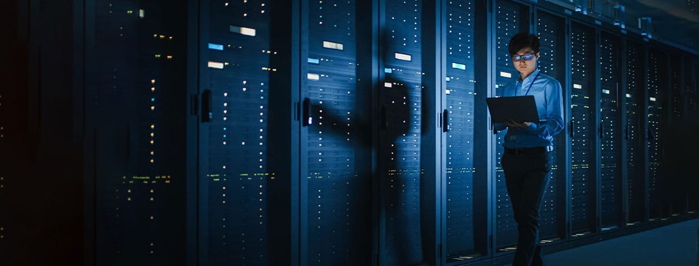 A person stands in a dimly lit server room, holding a laptop and inspecting rows of illuminated server racks.