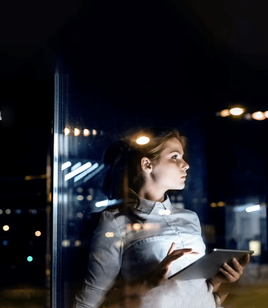 A woman holding a tablet stands by a window at night, looking outside with city lights reflected in the glass.