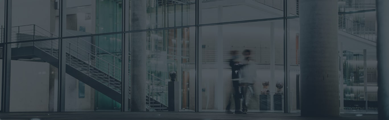Two blurred figures stand outside a modern glass building with a visible staircase and interior lights at night.