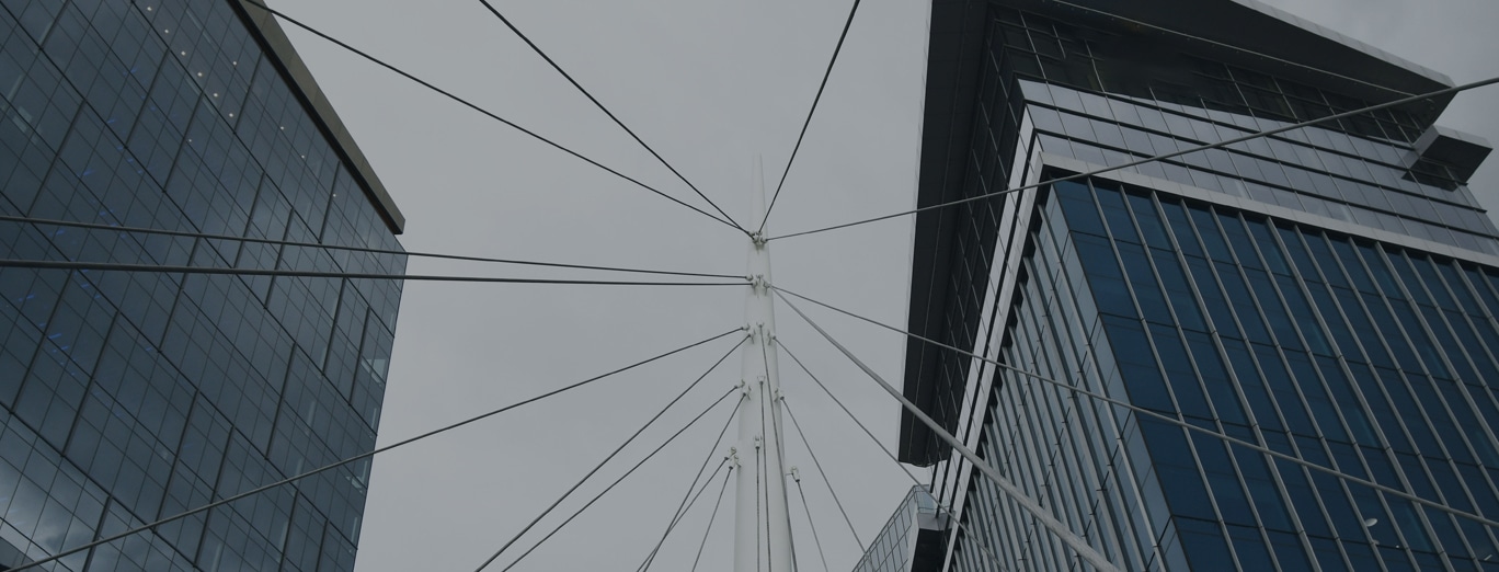 View looking up at the cables of a modern suspension bridge between two glass office buildings against a gray sky.