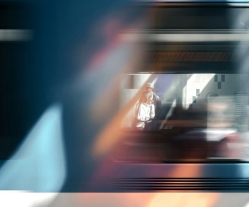 A person in a white shirt is seen through the window of a moving train, with blurred motion and light reflections in the foreground.