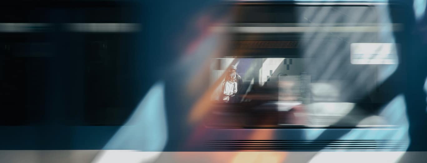 A blurred train moves past a woman standing on a platform, visible through a window, with reflections and light patterns on the glass.