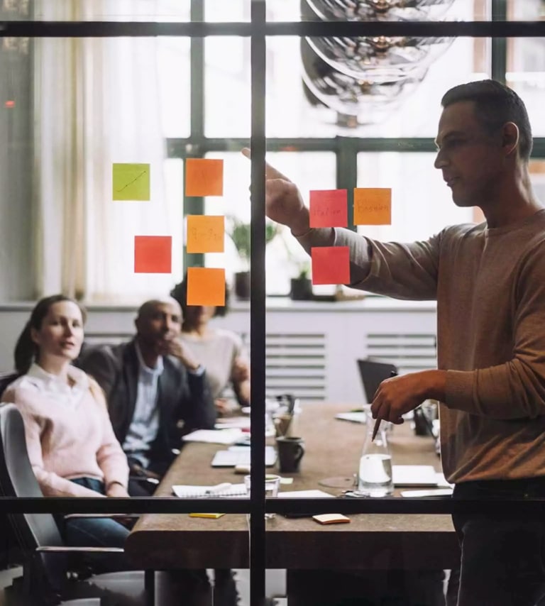 A person stands by a glass wall with colorful sticky notes, presenting to three seated colleagues in a modern meeting room.