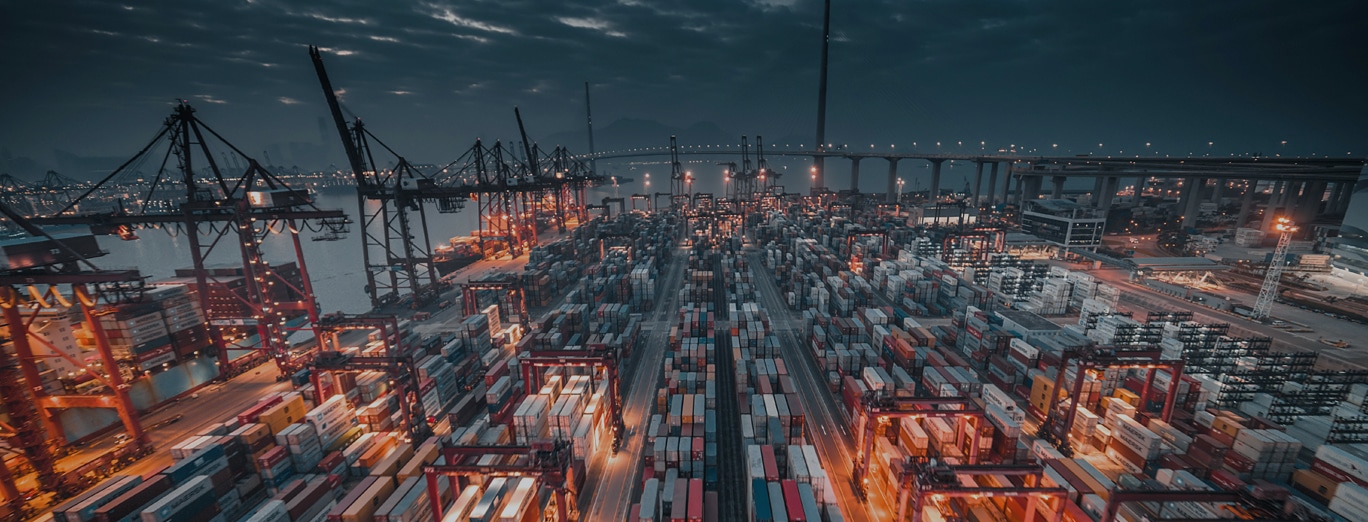 Aerial view of a busy shipping port at night, with numerous shipping containers, cranes, and illuminated lights under a cloudy sky.