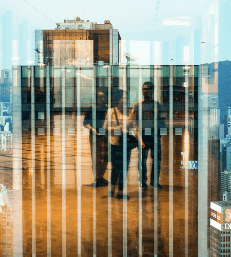 Reflection of three people standing indoors is visible in a glass window, with a cityscape and tall buildings faintly seen through the glass.