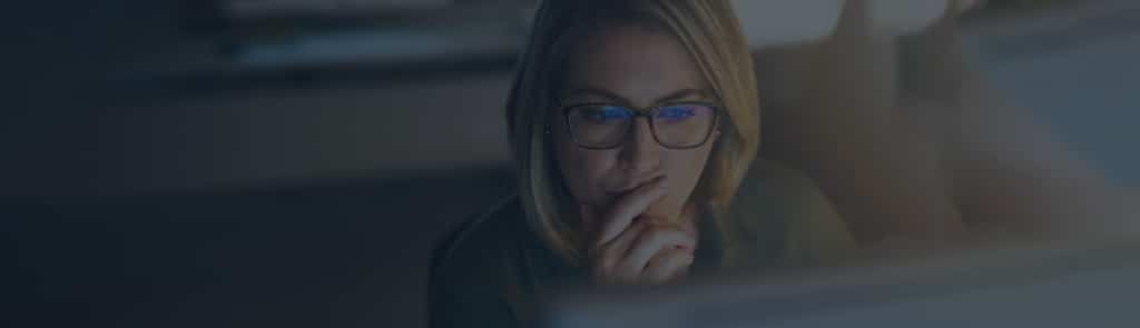 A woman wearing glasses looks at a computer screen with a thoughtful expression, resting her hand on her chin.