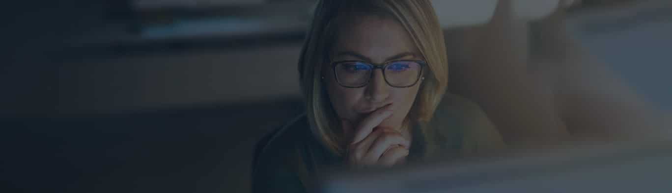 A woman wearing glasses looks at a computer screen with a thoughtful expression, resting her hand on her chin.