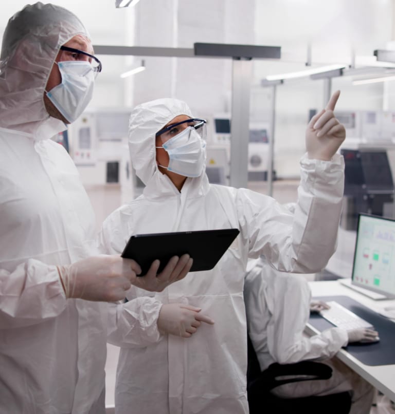 Two people in protective suits and masks discuss while one holds a tablet and points. Another person works at a computer in a clean, high-tech laboratory environment.
