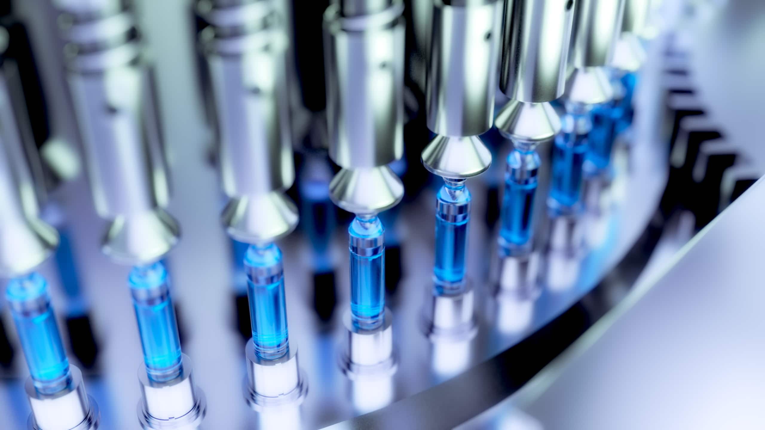 Automated machinery fills glass vials with blue liquid in a pharmaceutical manufacturing process.