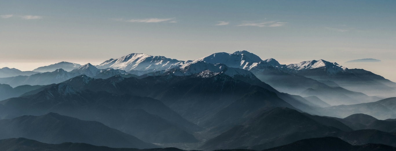 Distant snow-capped mountains rise above layers of dark, misty ridges under a hazy sky.