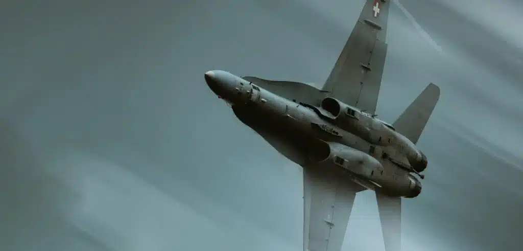 A military jet flies at an angle through a cloudy sky, displaying its underside with visible landing gear doors and national insignia on the vertical stabilizer.