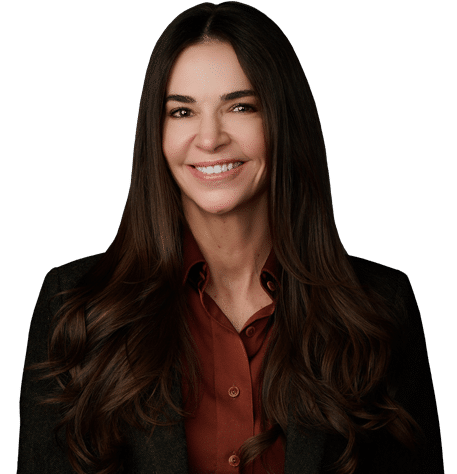 Woman with long brown hair wearing a dark blazer over a brown button-up shirt, smiling and facing the camera against a plain white background.