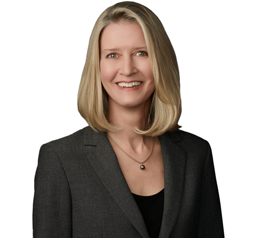 A woman with straight blond hair wearing a dark blazer, black top, and a necklace, smiling in a professional headshot against a plain background.
