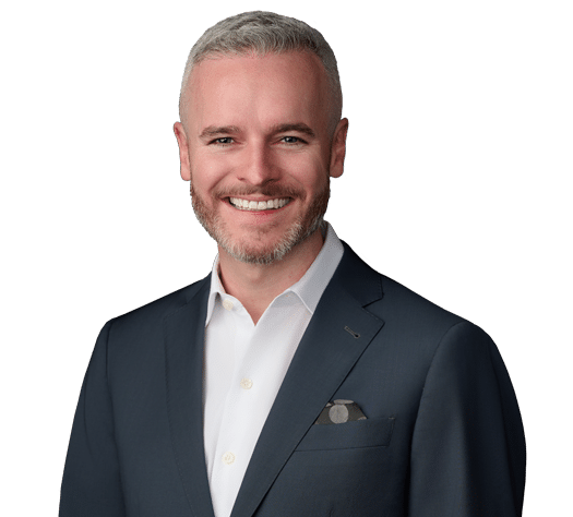Smiling man with short gray hair and beard wearing a dark suit jacket and white dress shirt, standing against a plain light background.