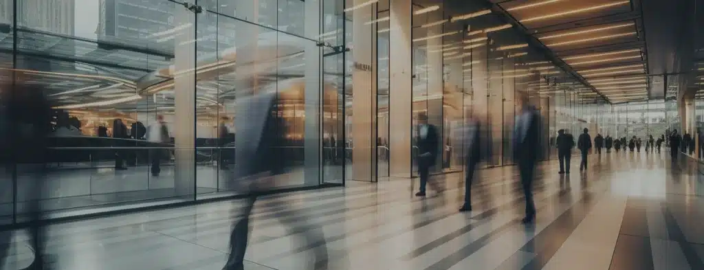 Blurred figures walk past a modern glass building entrance, with people and office lights visible inside. The scene suggests an urban business environment.