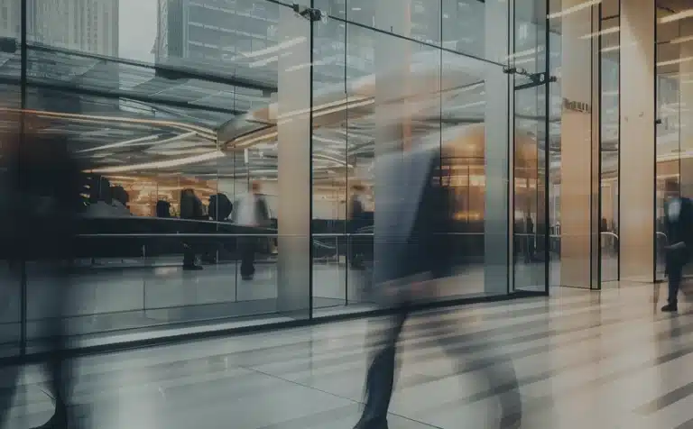 Blurred figures walk past a modern glass building entrance, with people and office lights visible inside. The scene suggests an urban business environment.