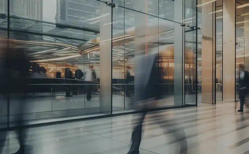 Blurred figures walk past a modern glass building entrance, with people and office lights visible inside. The scene suggests an urban business environment.