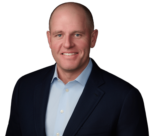 A man with a shaved head wearing a dark suit jacket and light blue dress shirt, smiling at the camera against a plain white background.