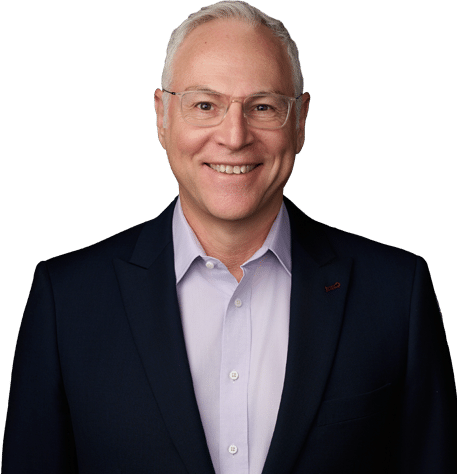 A middle-aged man with short gray hair and glasses, wearing a dark suit jacket over a light purple shirt, smiling at the camera against a plain white background.