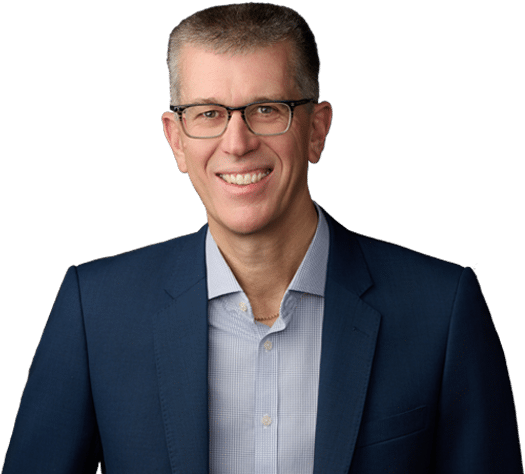 A man wearing glasses, a blue suit jacket, and a light-colored shirt is smiling at the camera against a plain white background.