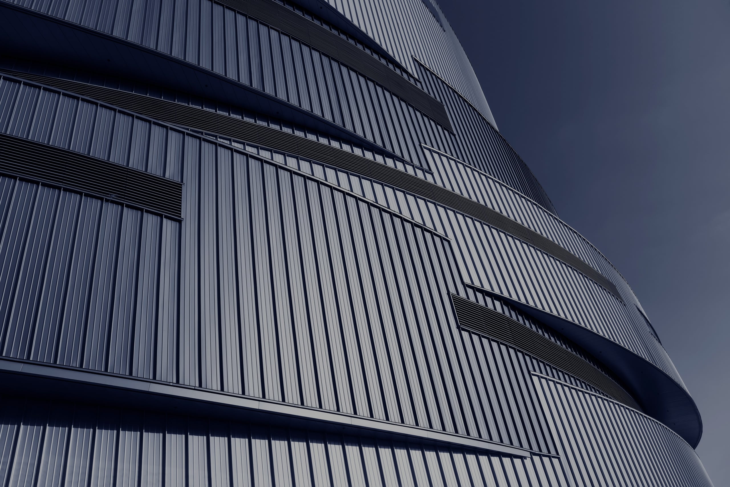 Close-up view of a modern curved building facade with overlapping, vertically ridged metal panels and narrow horizontal vents.