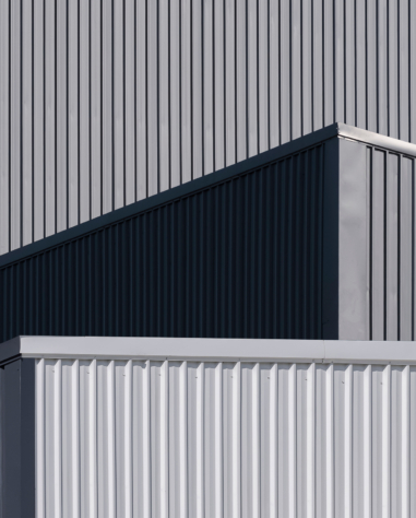 A close-up view of the exterior of a modern industrial building with overlapping geometric shapes and vertical metal siding in shades of grey.