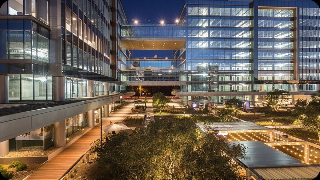 Modern glass office buildings with illuminated windows and outdoor courtyard, photographed at dusk.