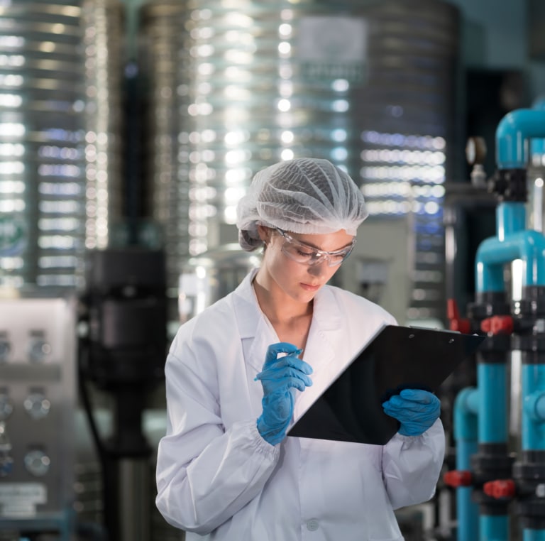 A person in a lab coat, hair net, safety glasses, and gloves writes on a clipboard in an industrial or laboratory setting with large metal tanks and pipes in the background.