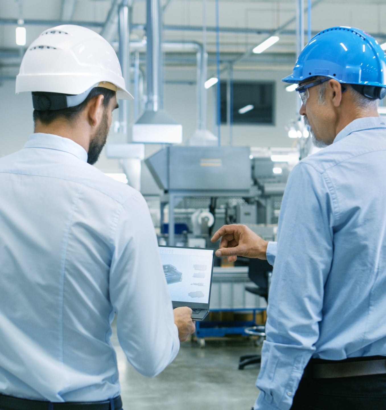 Two men in hard hats and dress shirts review information on a tablet in an industrial facility with machinery in the background.