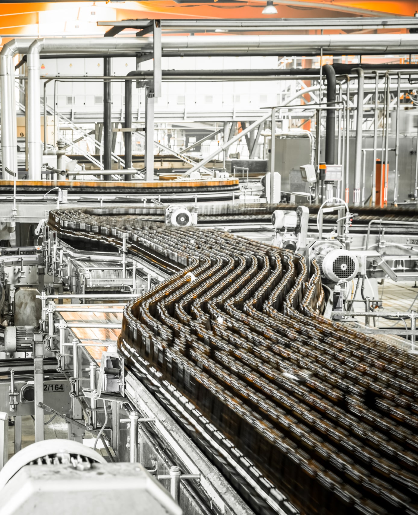 Automated conveyor system in a factory transports rows of empty glass bottles through a production line surrounded by industrial machinery and pipes.