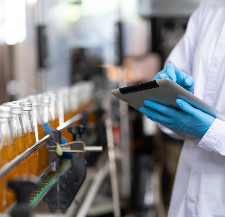 Person in a lab coat and blue gloves inspects a production line of filled glass bottles, using a tablet for data entry or quality control.