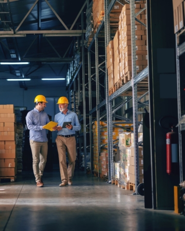 Two workers wearing yellow hard hats walk and review documents in a warehouse aisle lined with shelves and boxes.