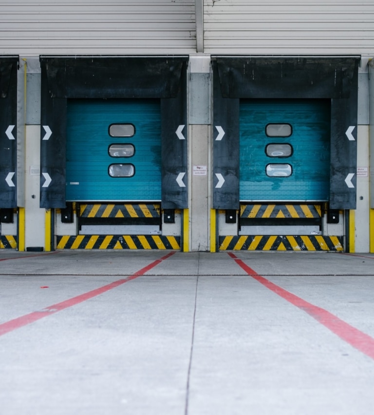 Two closed blue loading dock doors with black bumpers and yellow-black warning stripes at a warehouse, viewed from the concrete driveway marked with red lines.