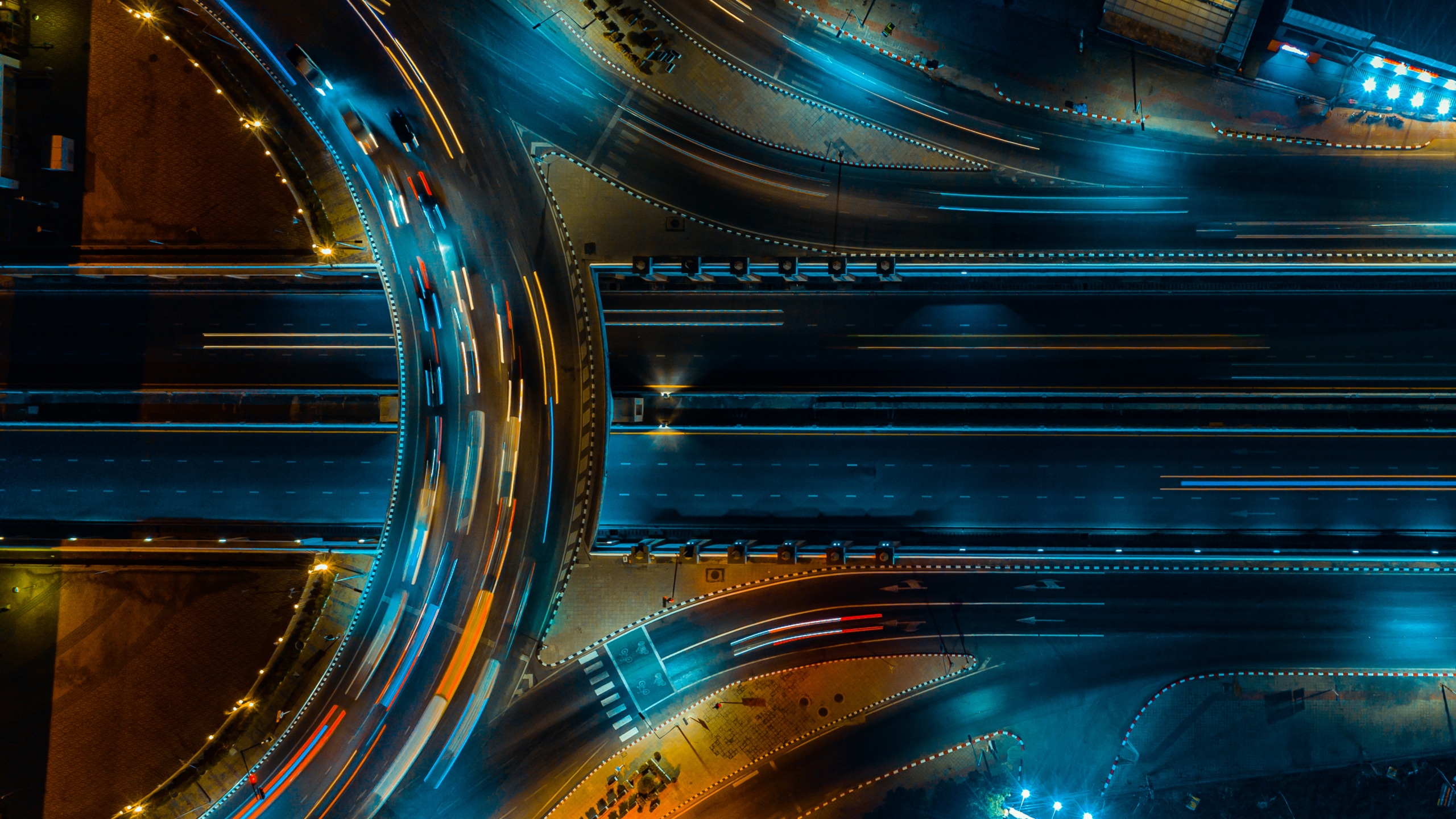 Aerial view of a highway interchange at night with cars creating light trails; streets are illuminated by blue and yellow lights.