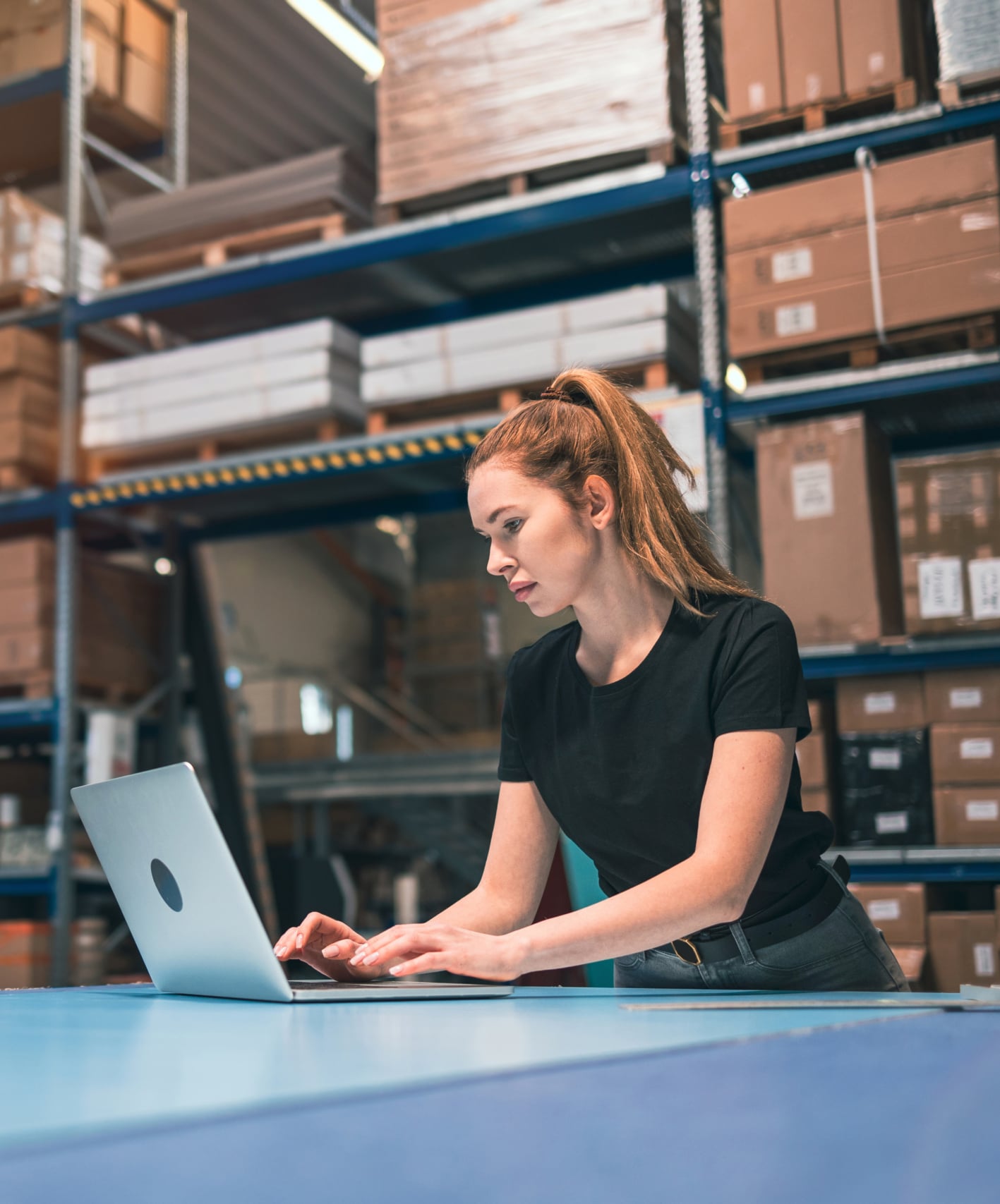 A woman in a black shirt uses a laptop on a blue table in a warehouse with shelves stacked with cardboard boxes in the background.