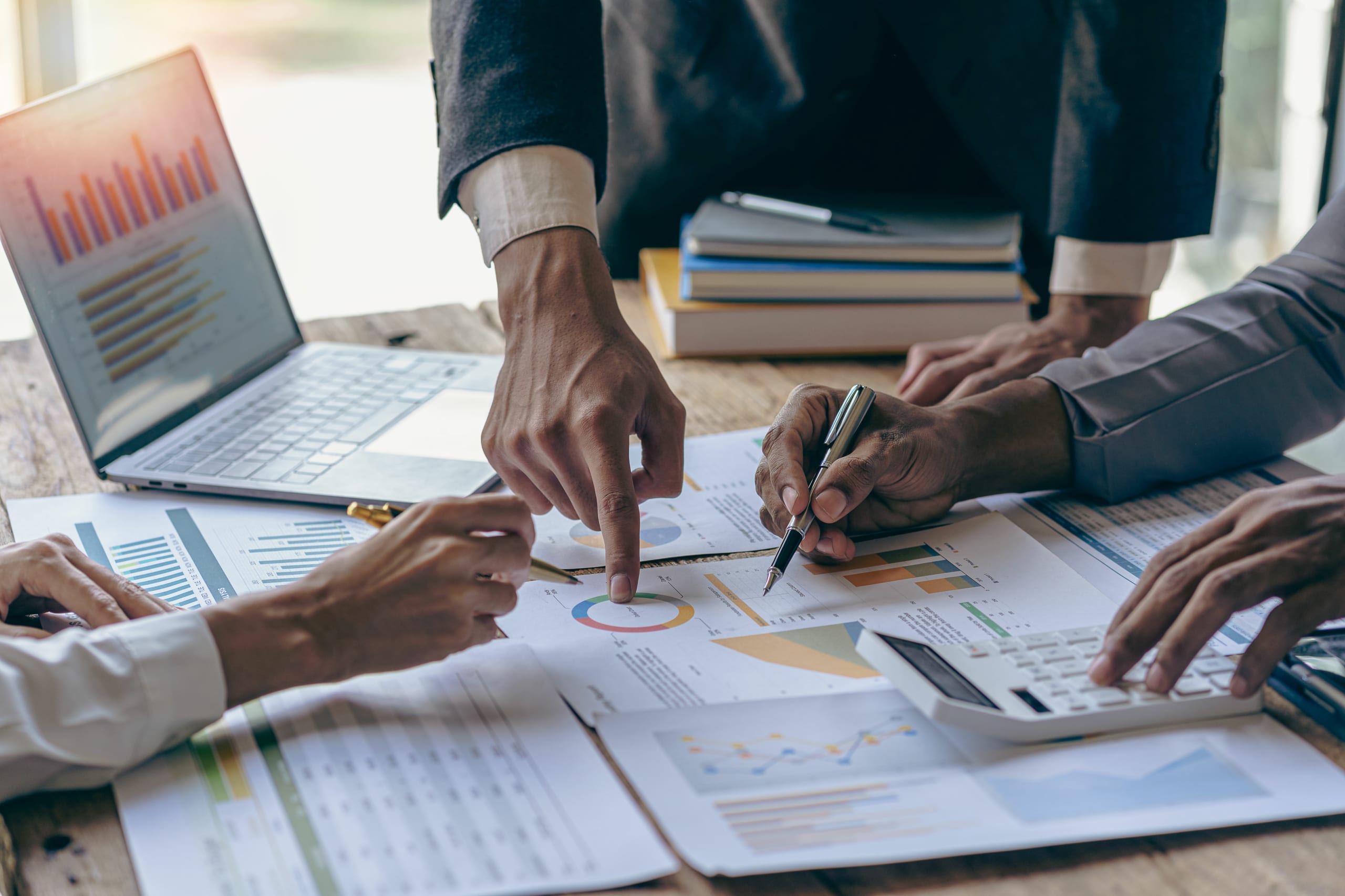 Three people review financial charts, graphs, and documents at a table, with one person pointing at a printed report and another using a calculator. A laptop is open nearby.