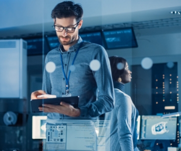 A man in an office setting looks at a tablet while a woman stands behind him, both surrounded by computer monitors and technical equipment.