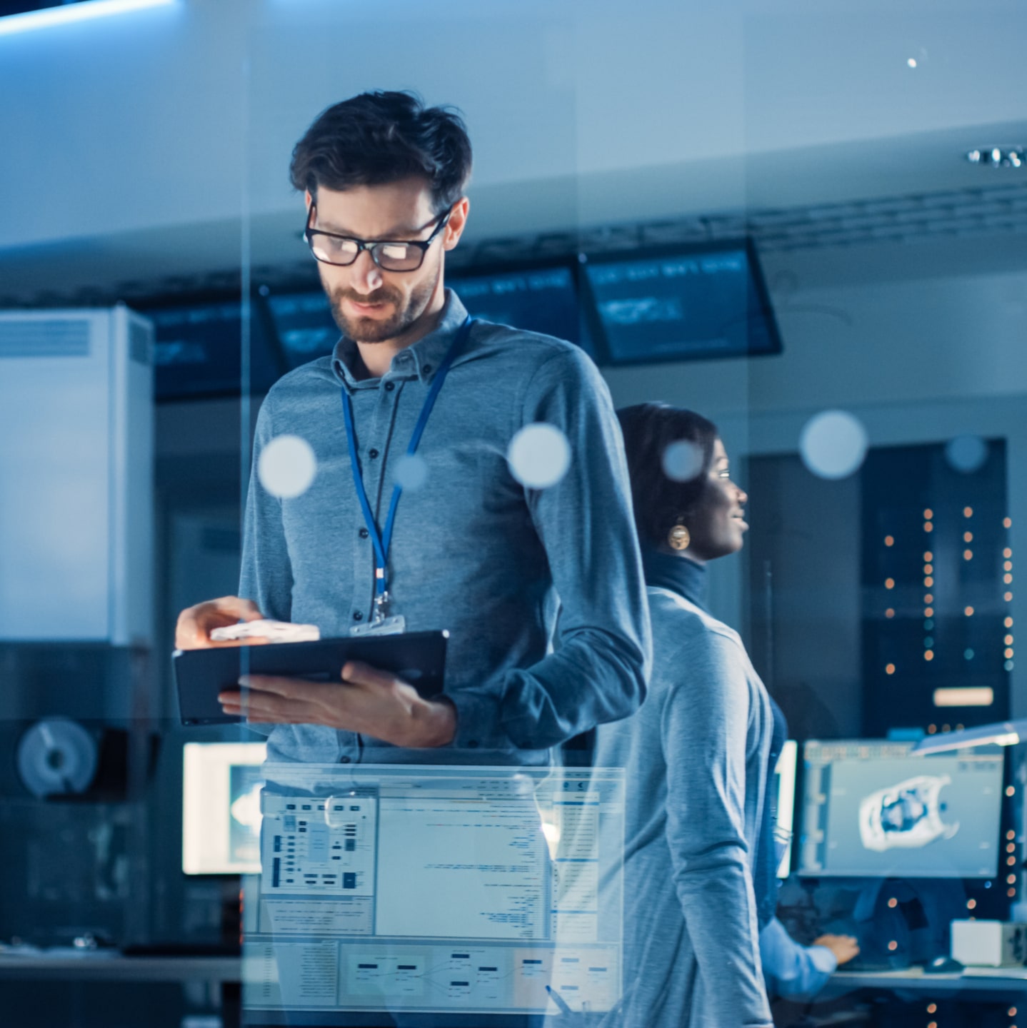 A man in an office setting looks at a tablet while a woman stands behind him, both surrounded by computer monitors and technical equipment.