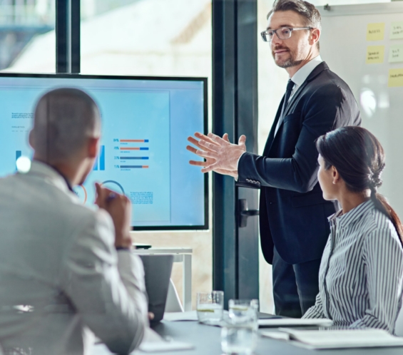 A man in a suit gives a presentation to two colleagues in an office, displaying a bar chart on a large monitor.