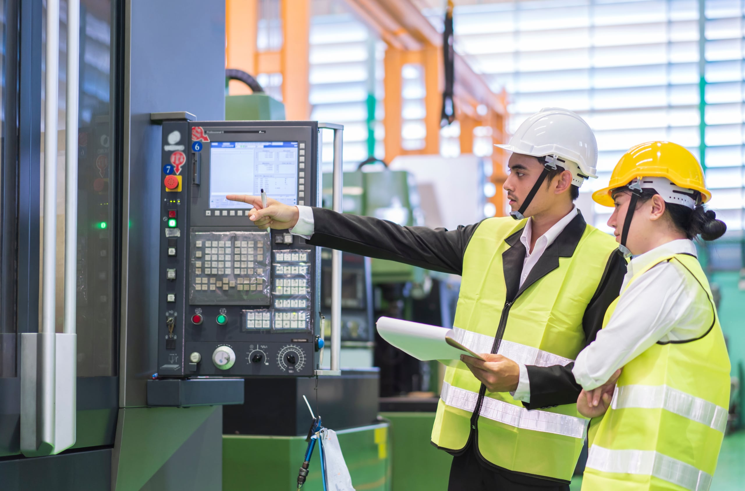 Two workers in safety helmets and vests operate and discuss settings on an industrial control panel in a factory environment.