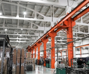 Spacious factory interior with tall orange columns, machinery, stacked pallets, and bright overhead lighting.