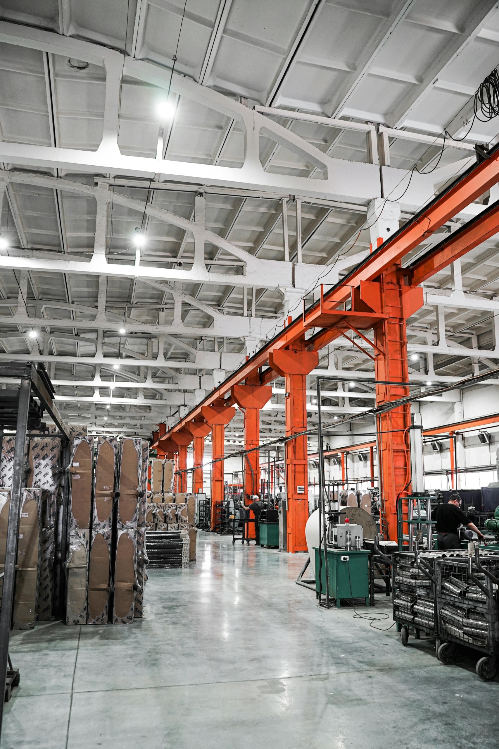 Spacious factory interior with tall orange columns, machinery, stacked pallets, and bright overhead lighting.