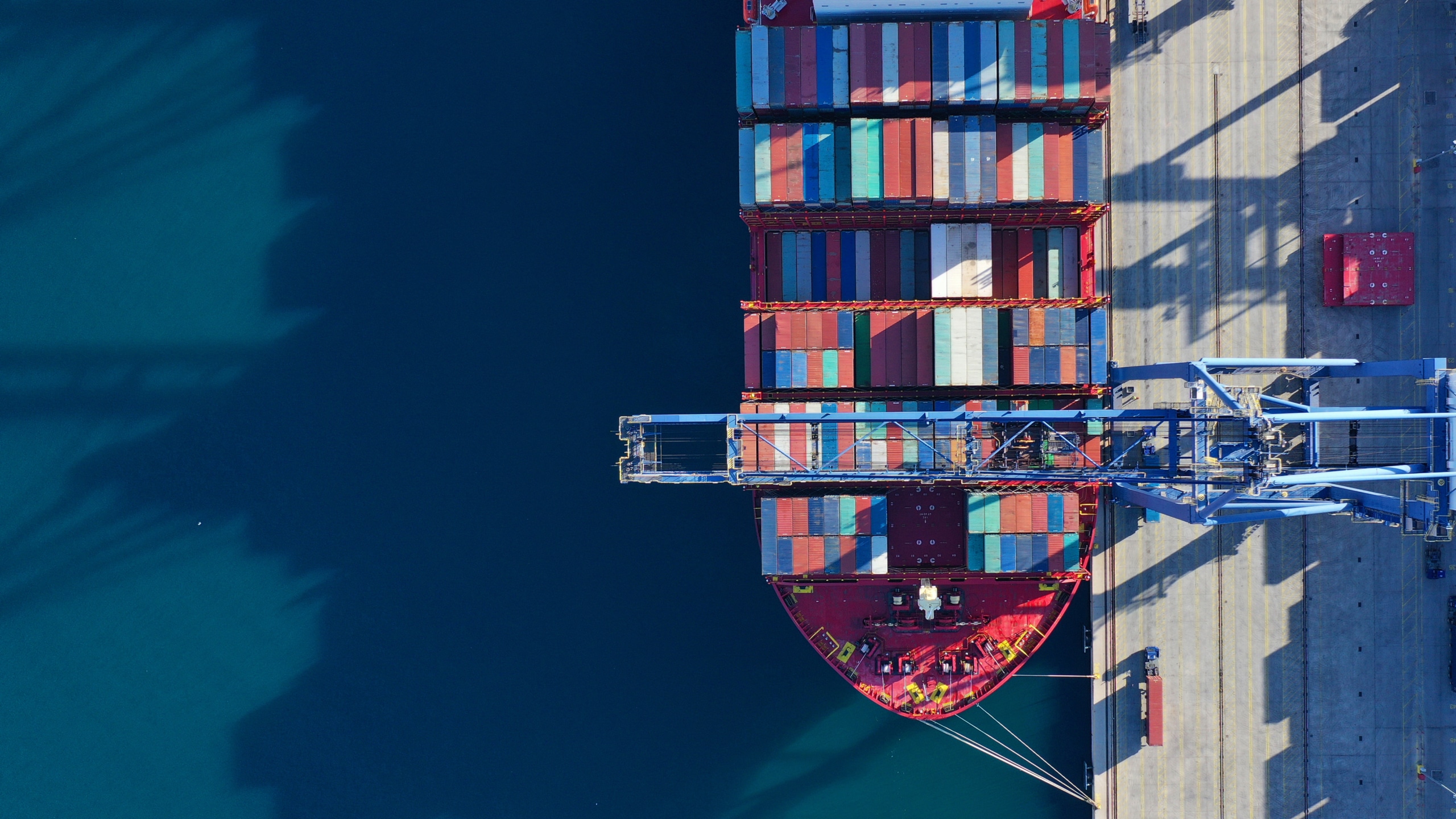 Aerial view of a cargo ship docked at a port, loaded with colorful shipping containers, next to a large crane and clear blue water.