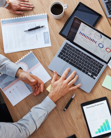 Two people review financial charts and marketing projections on paper and digital devices at a wooden desk with coffee, notebooks, and a laptop displaying graphs.